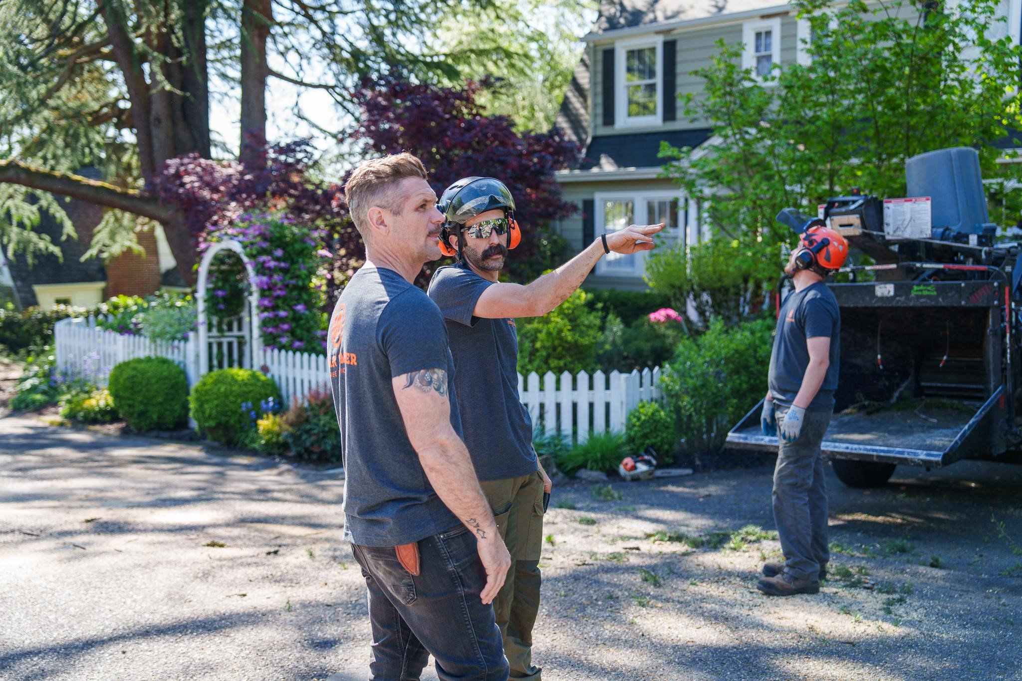 Two workers discussing while standing near a tree and a chipper truck.